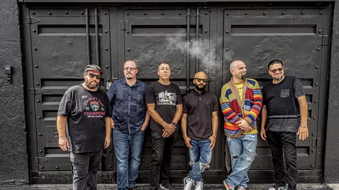 Portrait of a six-piece band leaning on a wall.