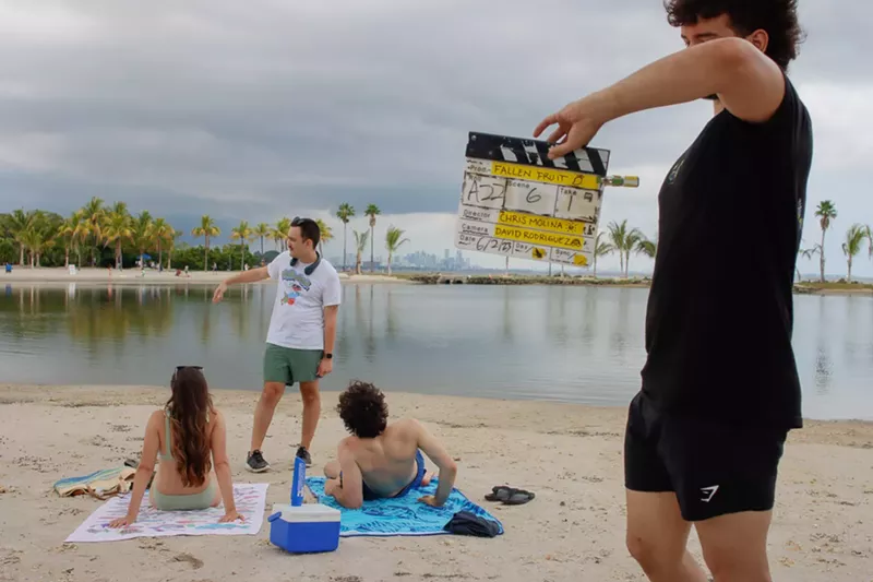 Image: Chris Molina is speaking with actors lying on the beach while a man holds a film slate in the foreground