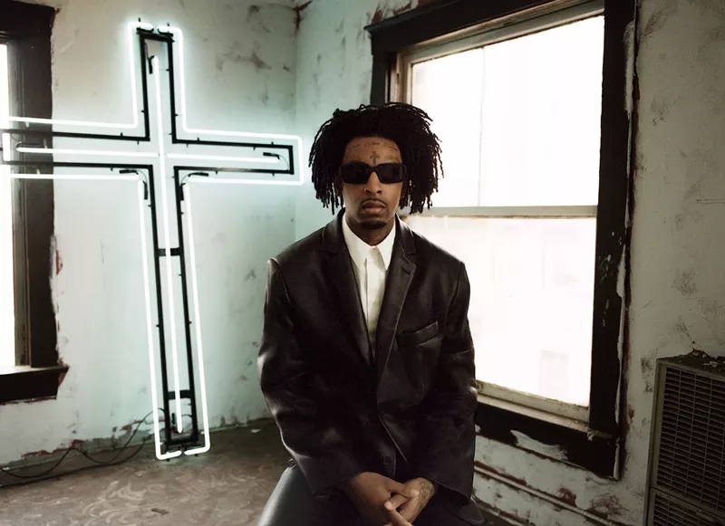 Image: 21 Savage sitting down with a cross behind him