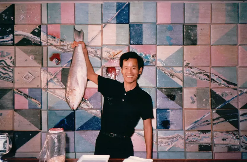 Image: vintage photo of a smiling man dressed in black, standing at a counter against a tiled background and holding up a whole fish in his right hand
