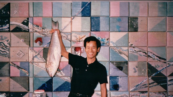 vintage photo of a smiling man dressed in black, standing at a counter against a tiled background and holding up a whole fish in his right hand