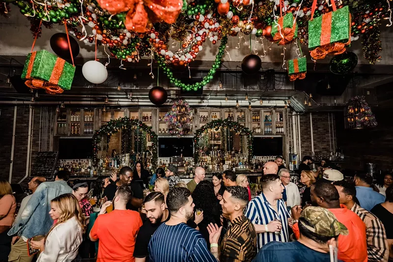 Image: Guests mingle with cheer as they're surrounded by fun holiday decorations at Bodega Taqueria y Tequila. The ceiling is draped in holiday decoras guests hold and sip colorful holiday cocktails.