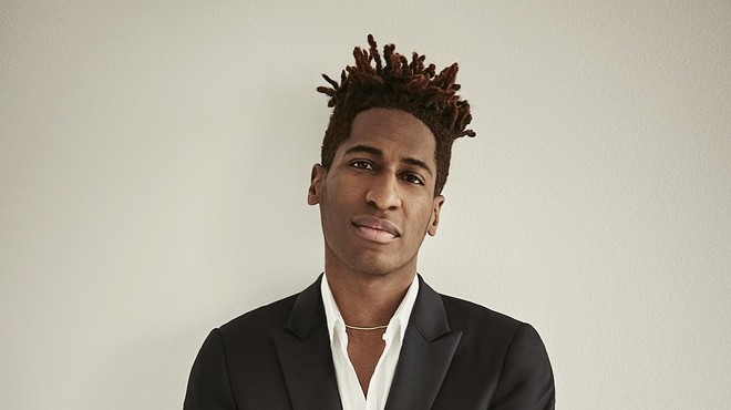 Jon Batiste in a suit standing against a beige background