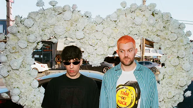 Two musicians are posing in front of a heart made out of flowers.