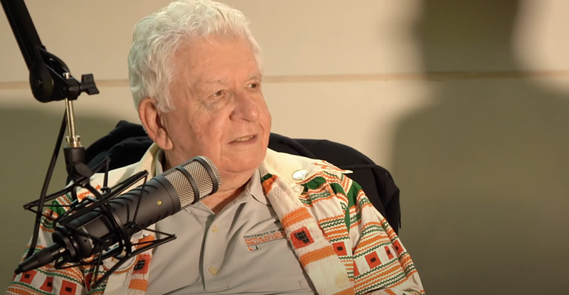 Image: An elderly man with white hair looks to the side in front of a podcast microphone