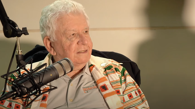 An elderly man with white hair looks to the side in front of a podcast microphone