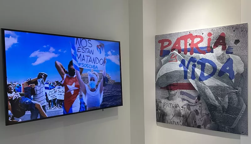 Image: A TV in a museum displays images of Cuban protesters. One holds up a sign reading, "Nos Estan Matando (They're killing us) SOS Cuba." On the right of the TV, a painting displays the slogan, "Patria y Vida (Homeland and Life)"