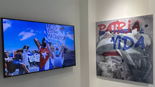 A TV in a museum displays images of Cuban protesters. One holds up a sign reading, "Nos Estan Matando (They're killing us) SOS Cuba." On the right of the TV, a painting displays the slogan, "Patria y Vida (Homeland and Life)"