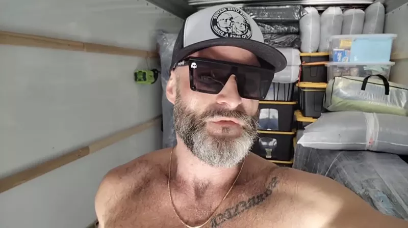 Image: a shirtless Jon Minadeo speaks into the camera inside a loaded U-Haul.