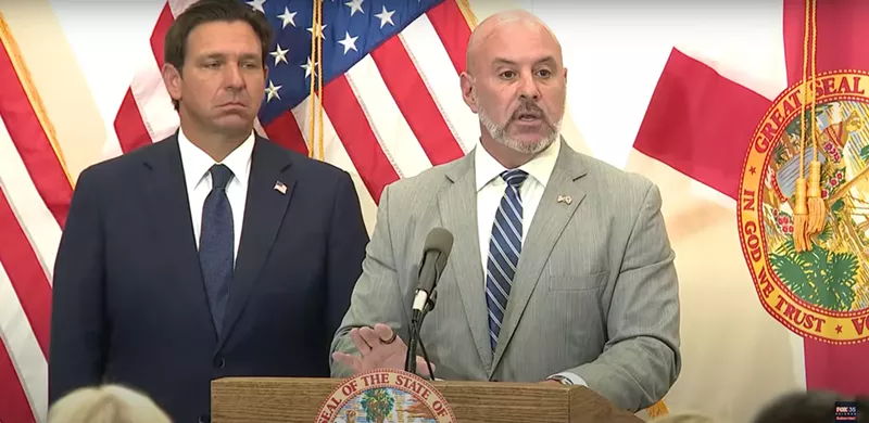Image: Blaise Ingoglia speaking at a press conference and Ron DeSantis stands to his left with a frown on his face.