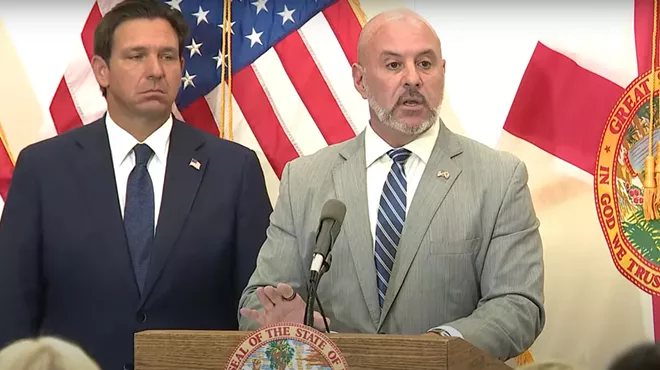 Blaise Ingoglia speaking at a press conference and Ron DeSantis stands to his left with a frown on his face.
