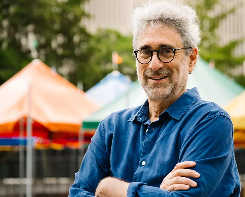 Image: Color photo of Mitchell Kaplan, founder of Books & Books, an independent bookstore chain based in Coral Gables (Miami), Florida. In this horizontal image, the bespectacled bookseller stands with arms crossed, in front of tents set up in downtown Miami for the annual Miami Book Fair, an event Kaplan cofounded in 1984.