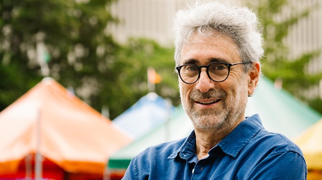 Color photo of Mitchell Kaplan, founder of Books & Books, an independent bookstore chain based in Coral Gables (Miami), Florida. In this horizontal image, the bespectacled bookseller stands with arms crossed, in front of tents set up in downtown Miami for the annual Miami Book Fair, an event Kaplan cofounded in 1984.
