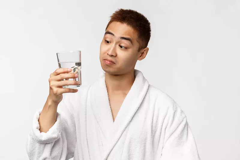 Image: Portrait of Asian man in bathrobe looking skeptical while holding glass of water
