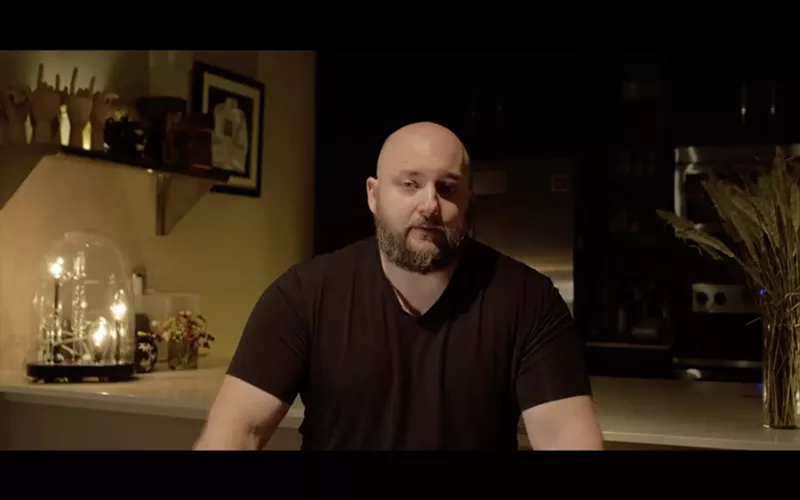 Image: screenshot of a man with a shaved head and a full beard, seated at a kitchen counter