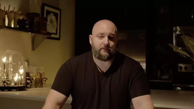 screenshot of a man with a shaved head and a full beard, seated at a kitchen counter