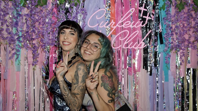Two women pose in front of a Curfew Club backdrop