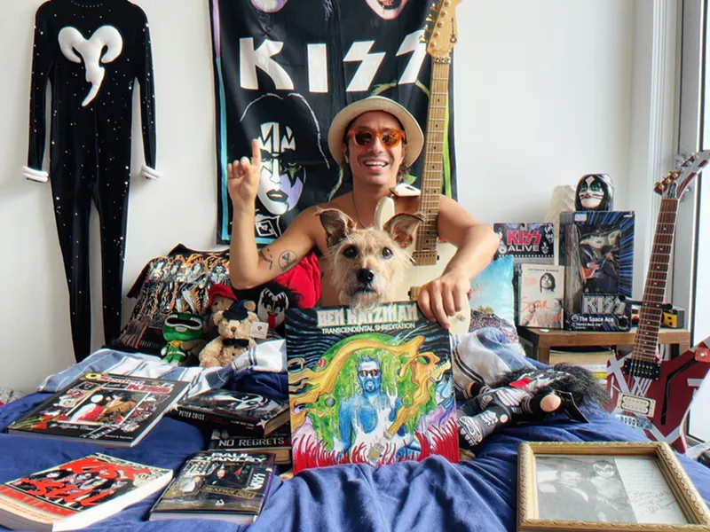 Image: Musician Ben Katzman sits on his bed holding a copy of his album, Transcendental Shredidation, with a Kiss poster hanging on his wall in the background