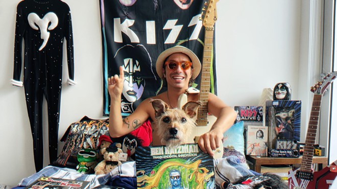 Musician Ben Katzman sits on his bed holding a copy of his album, Transcendental Shredidation, with a Kiss poster hanging on his wall in the background