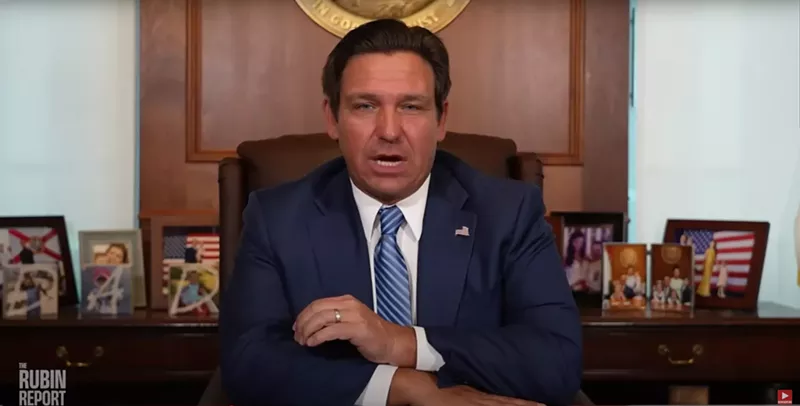 Image: Ron DeSantis wearing a blue tie sitting at his desk
