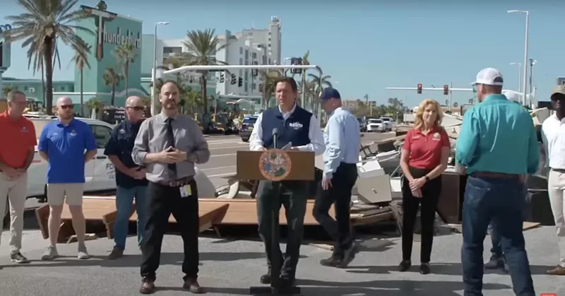 Image: Ron DeSantis stands in front of a trash pile during a press conference.
