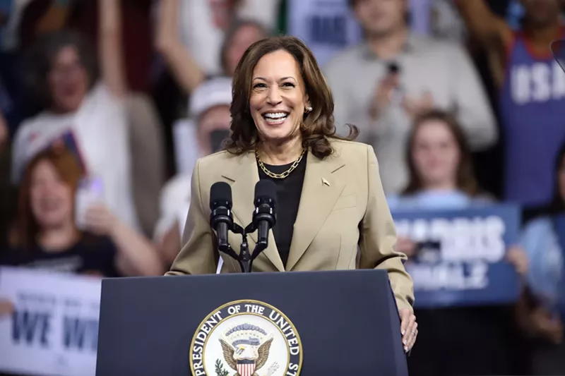 Image: photo of Kamala Harris smiling widely while standing at a lectern bearing the presidential seal, with a crowd of supporters in the background