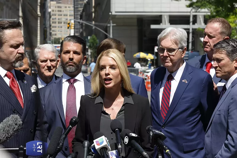 Image: Blond woman in front of a bank of microphones with several people around her, including Donald Trump Jr.