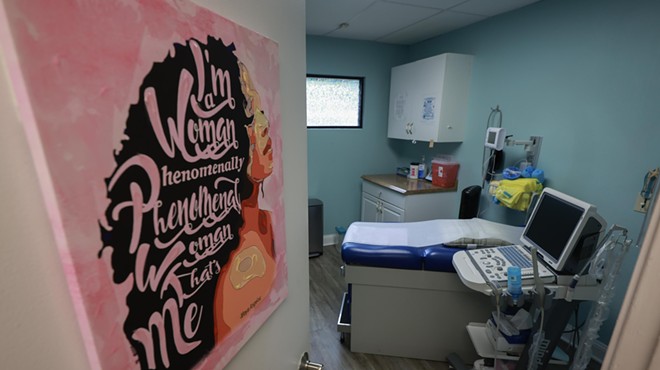 A door with a pink poster is propped open, leading to an examination room in an abortion clinic