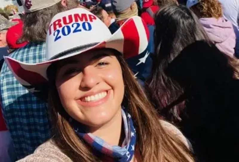 Image: color photo of a smiling young woman wearing a white, red, and blue Trump 2020 hat