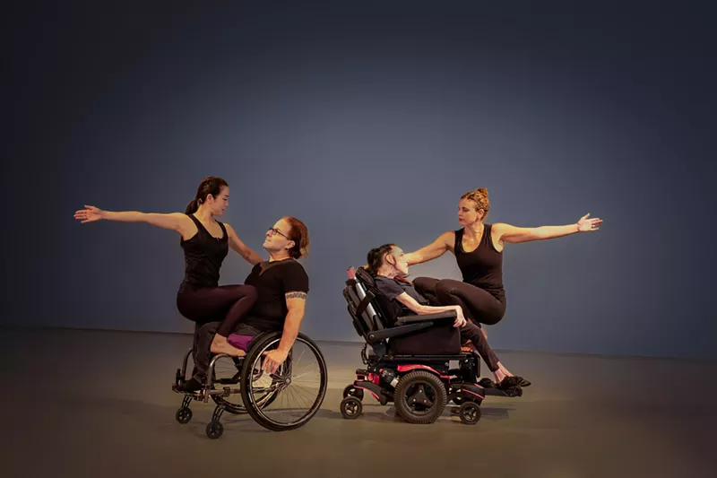 Image: color photo of two sets of partners performing a dance on a stage; each partnership consists of one dancer who uses a wheelchair and one who does not