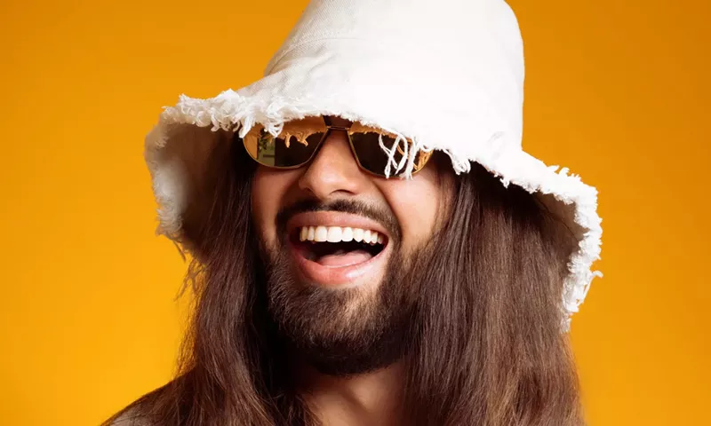 Image: Picture of a man with long hair wearing a white hat against a yellow background.
