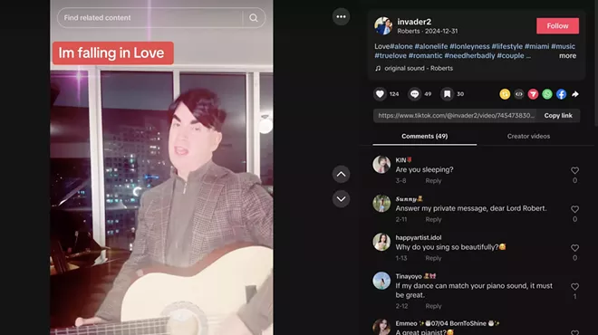 Screenshot of a man with dark eyebrows and a dark toupee singing and holding the acoustic guitar