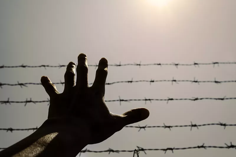 Image: A hand reaching before a barbed wire fence