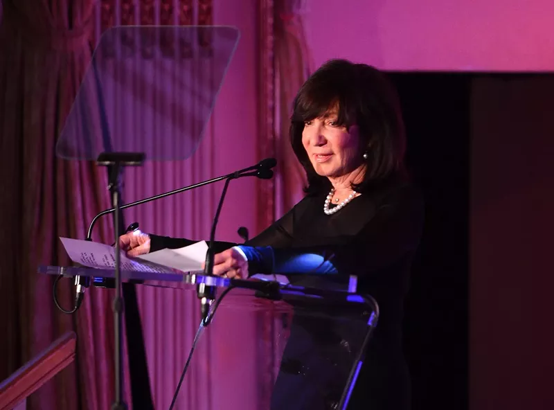 Image: photo of a woman standing at a podium and speaking into a microphone