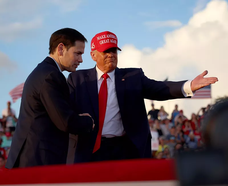 Image: Donald Trump in a "Make America Great Again" cap shakes Marco Rubio's hand in front of a large crowd while directing him to the podium at a Miami-Dade rally