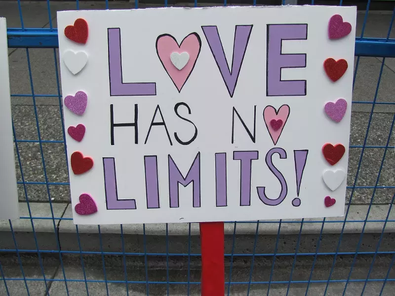 Image: a painted protest sign with heart stickers reads, "Love has no limits!"