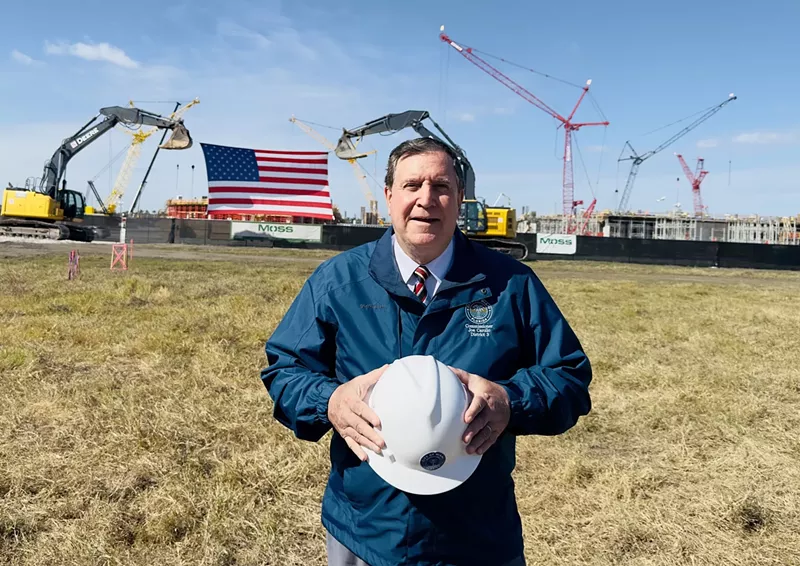 Image: photo of Miami City Commissioner Joe Carollo, wearing a jacket and holding a hardhat in a vacant field with cranes and earth moving machinery in the background