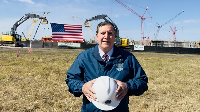 photo of Miami City Commissioner Joe Carollo, wearing a jacket and holding a hardhat in a vacant field with cranes and earth moving machinery in the background