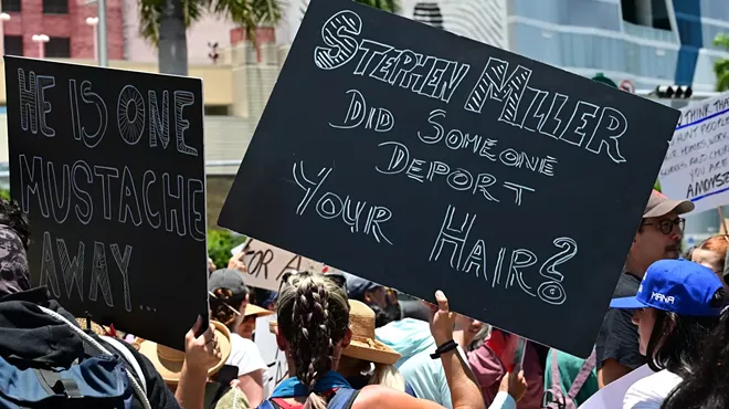 A protester with her back towards the camera holds a black sign that reads, "Stephen Miller did someone deport your hair?"