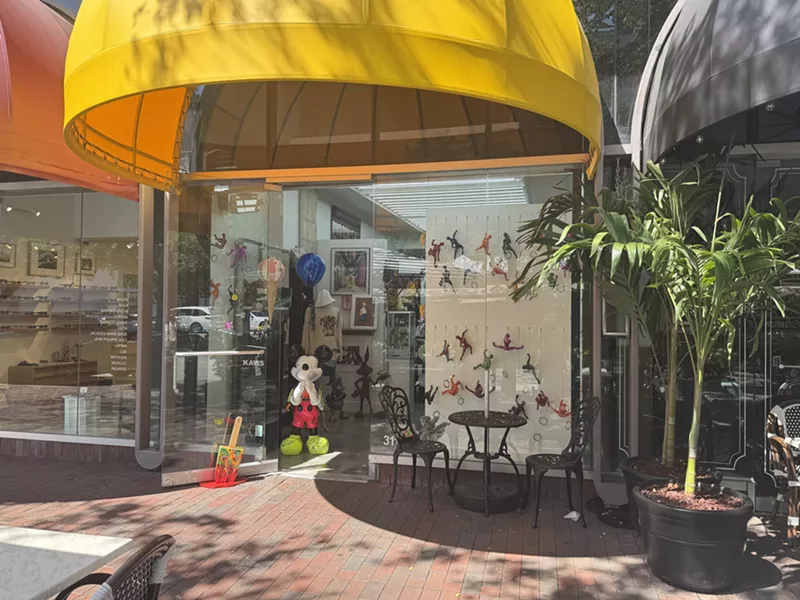 Image: Miami Art Gallery Gift Shop glass storefront, which has a Mickey Mouse sculpture at its entrance, along with a yellow awning.
