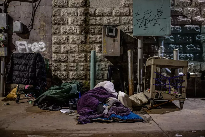 Image: A homeless person lies wrapped in a blanket on the concrete in front of a wall with the word, "Free," painted on it