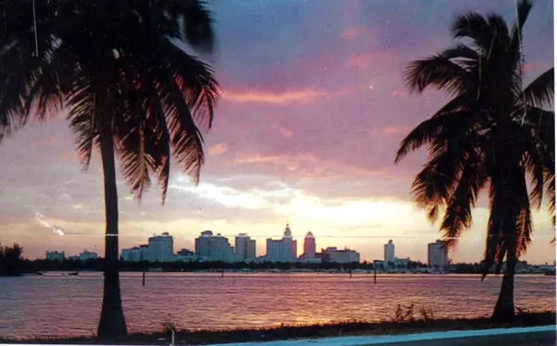 Image: color postcard from 1956 featuring a photo of the Miami skyline, flanked by two palm trees, with Biscayne Bay in the middle distance
