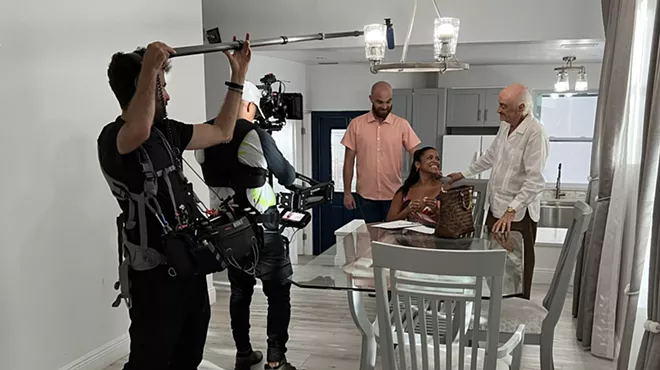 a camera operator and man with a boom mic record an acting scene with two men and a woman at a dining room table