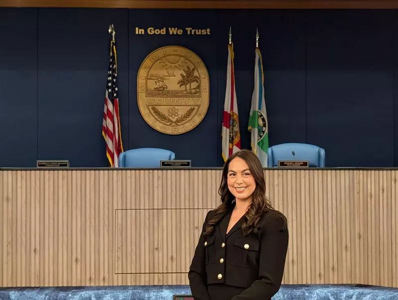Image: A brunette woman stands before a Miami-Dade County crest while wearing a black jacket