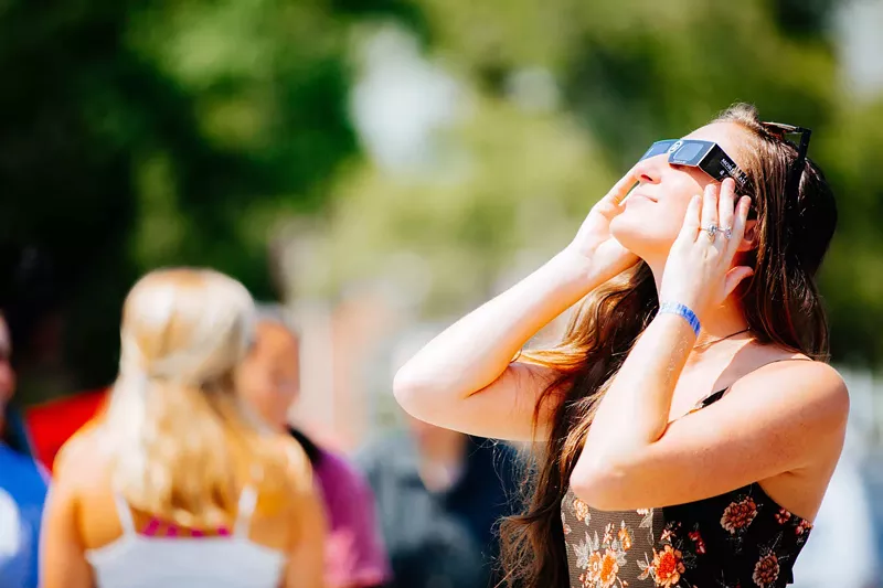 Image: Color photo of a young woman looks skyward during a solar eclipse while wearing the approved type of disposable eye protection