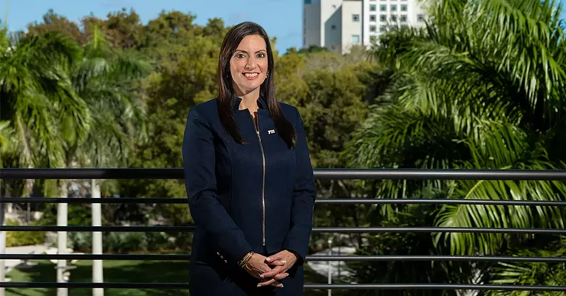Image: photo of a woman dressed in navy blue, standing with her hands clasped in front of her with tropical greenery and an FIU highrise in the background