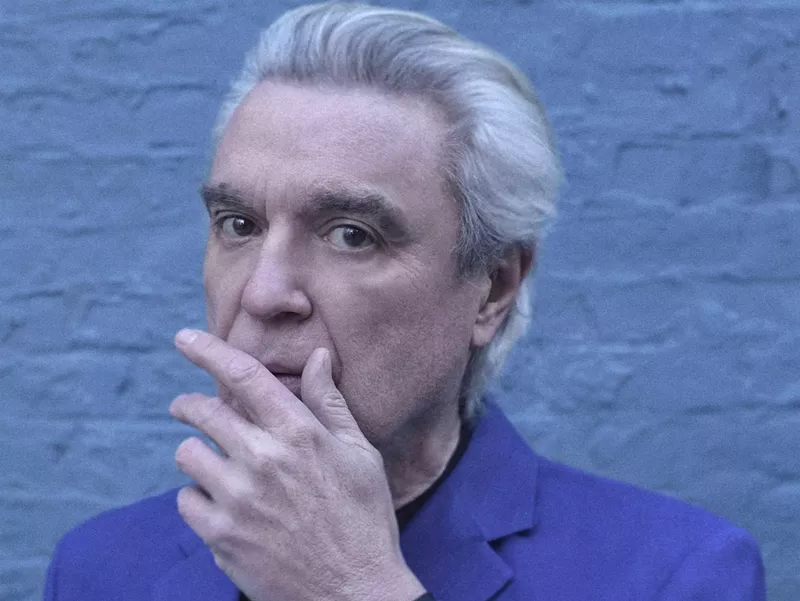 Image: cyan-tinged headshot of David Byrne, with his gray hair swept back and his left hand partially covering his mouth, as if stifling a cough