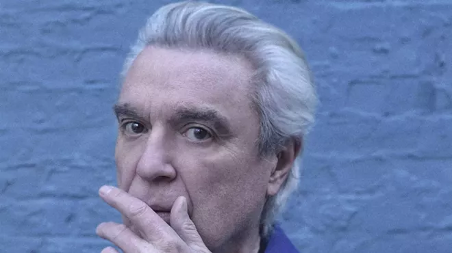 cyan-tinged headshot of David Byrne, with his gray hair swept back and his left hand partially covering his mouth, as if stifling a cough