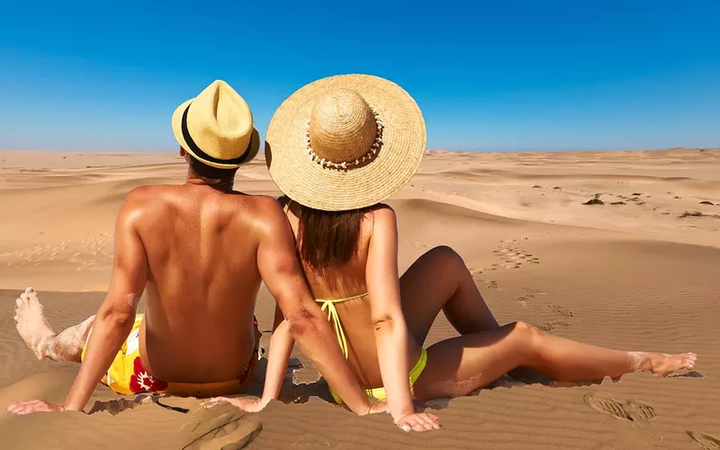 Image: photo-illustration depicting a bathing suit clad couple avec straw hats appearing to sunbathe in a Gambian desert as opposed to on tropical a beach