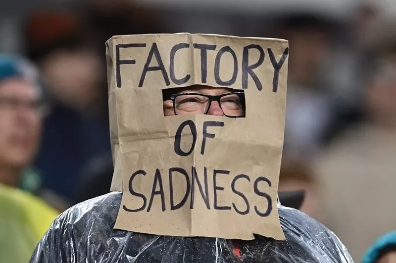 Image: football fan with brown paper bag on head that says "FACTORY OF SADNESS," written in all caps with a Sharpie